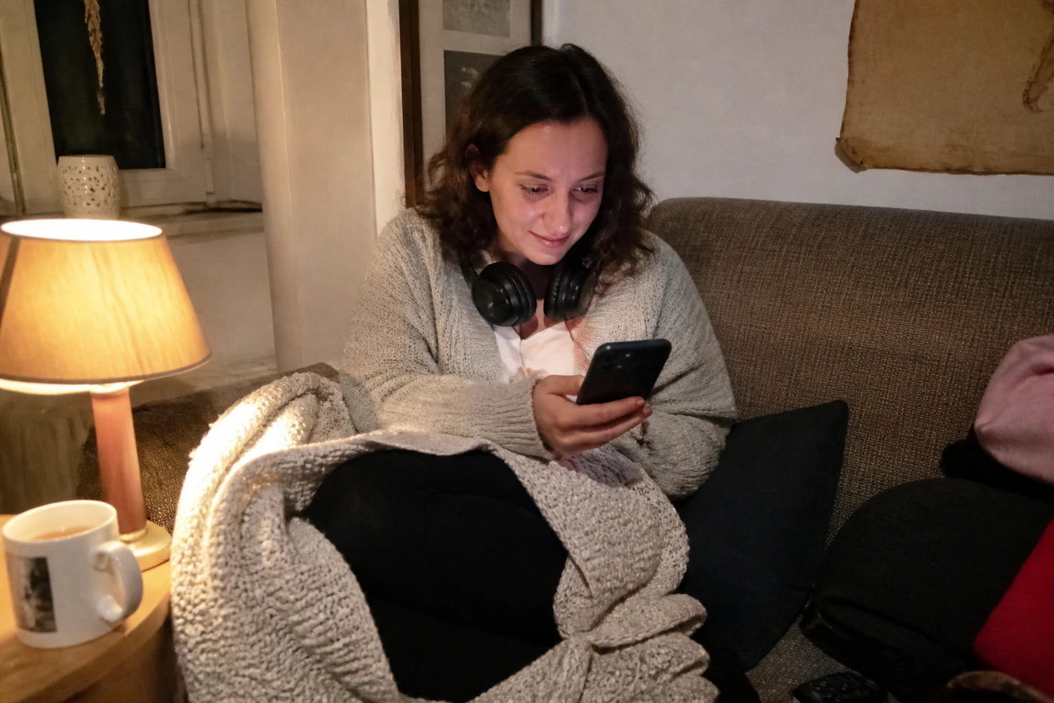 Abend-Schnappschuss: Frau mit Kopfhörern um den Hals sitzt eingekuschelt auf dem Sofa und lächelt beim Beantworten von Fragen auf ihrem Handy