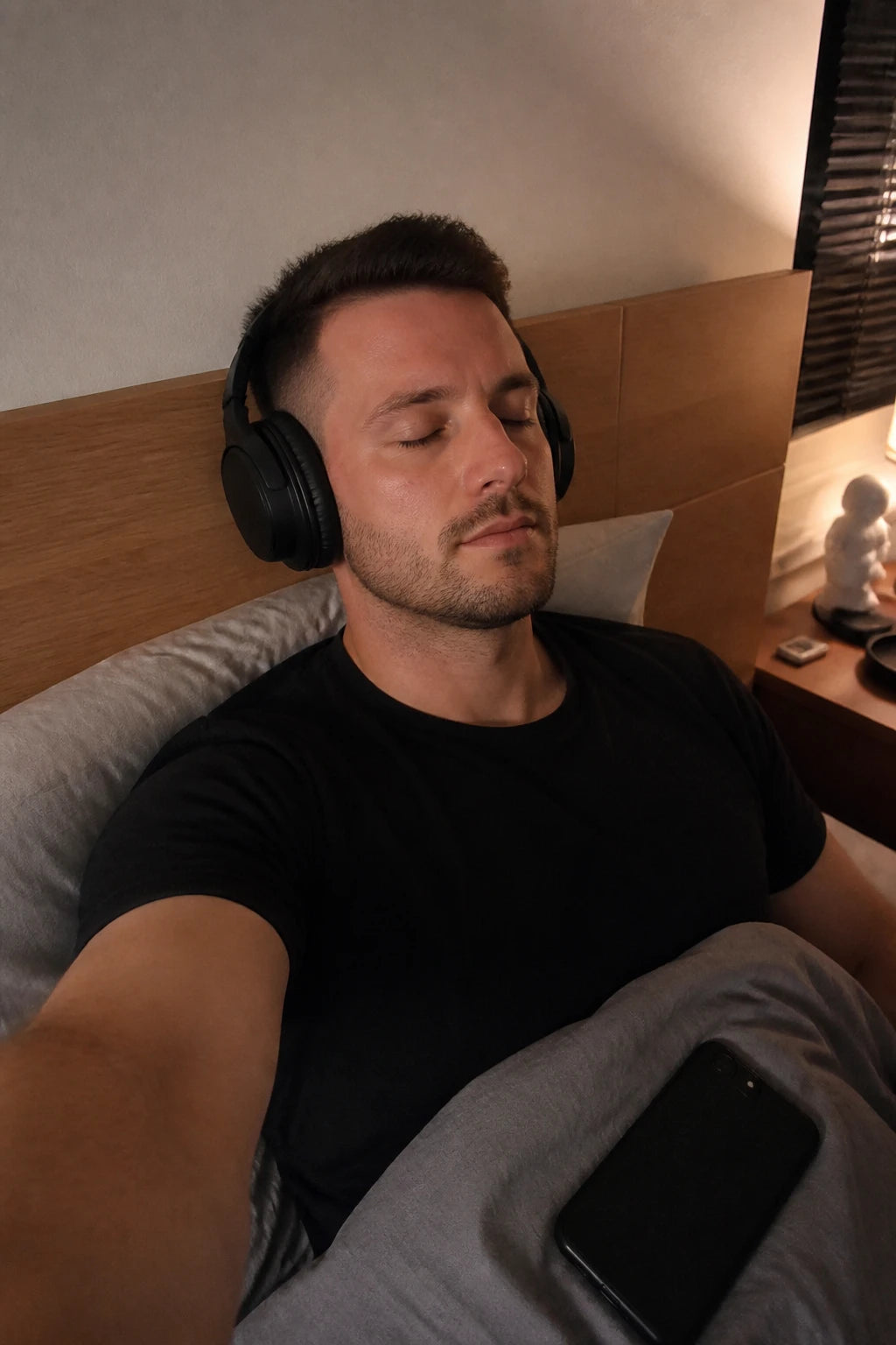 Selfie desselben Mannes im Bett: Kopfhörer auf, Augen zu, Handy umgedreht auf der Decke