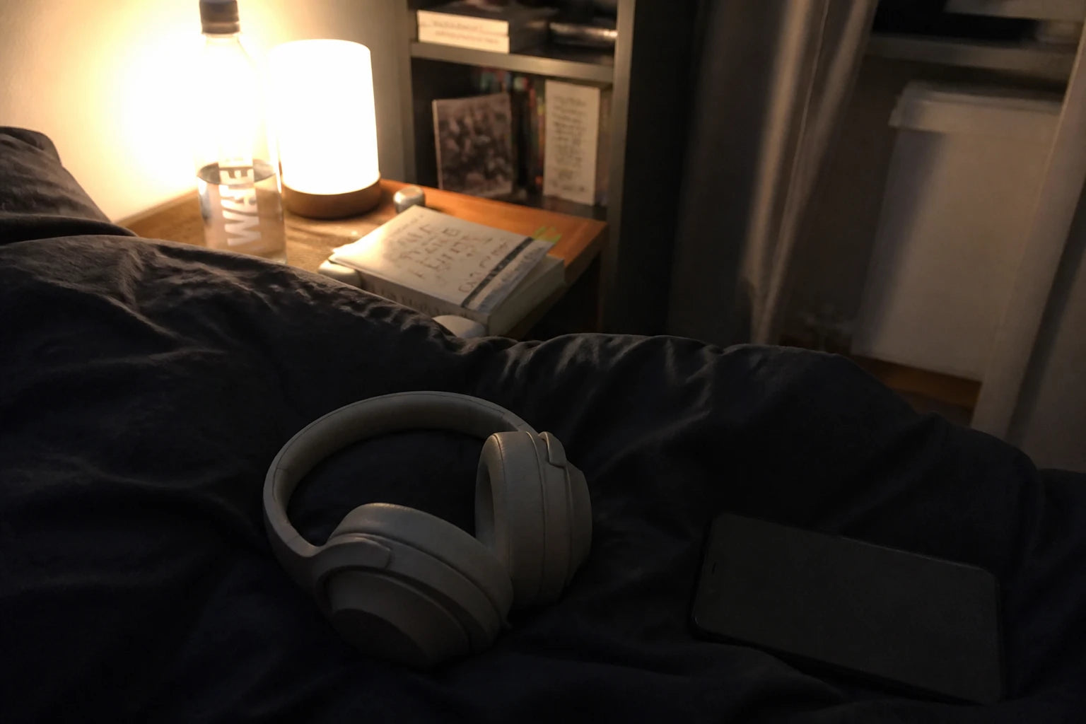Nachttisch bei Nacht: Kopfhörer auf der Bettdecke, Handy mit dem Display nach unten, warme Lampe