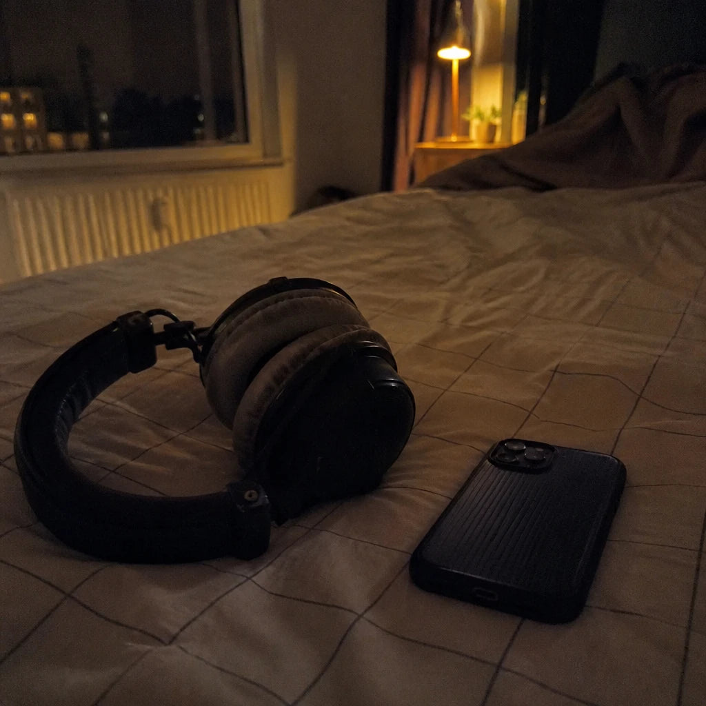 Kopfhörer auf dem Bett neben einem umgedrehten Handy bei Nacht, warme Lampe